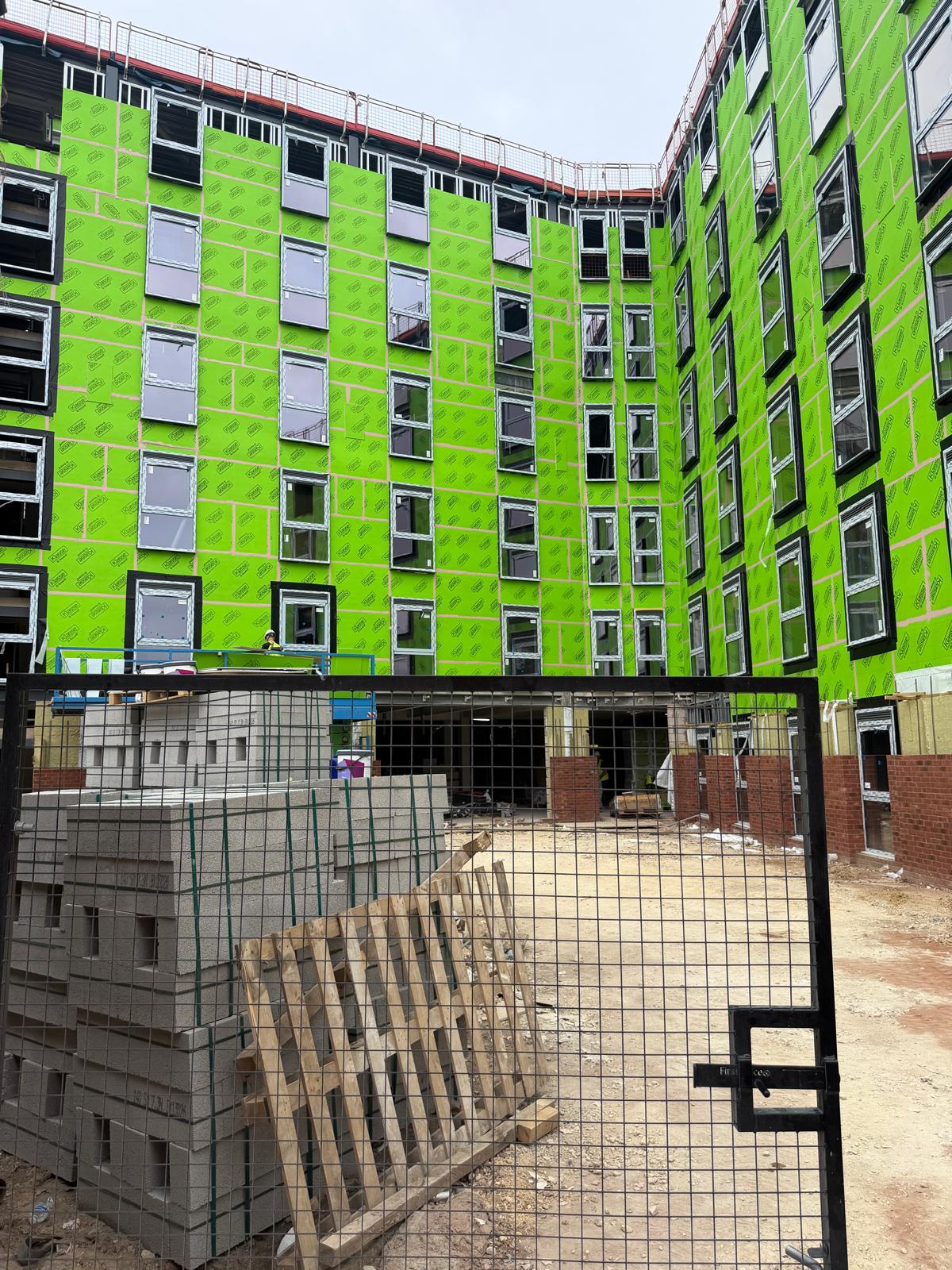 A construction site courtyard with a large building facade wrapped in bright green panels, viewed behind a black metal gate. Concrete blocks and wooden pallets are stacked in the foreground, and the ground is bare dirt.